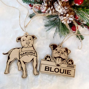 K&ouml;nnte beinhalten: Zwei Hundeornamente aus Holz mit eingravierten Details. Eines zeigt einen stehenden Hund mit dem Namen "SAKURA" auf einem Banner. Das andere zeigt einen Hund mit dem Namen "BLOUIE" und dem Jahr "2020" auf einem Schild. Beide haben Schnuraufh&auml;nger.