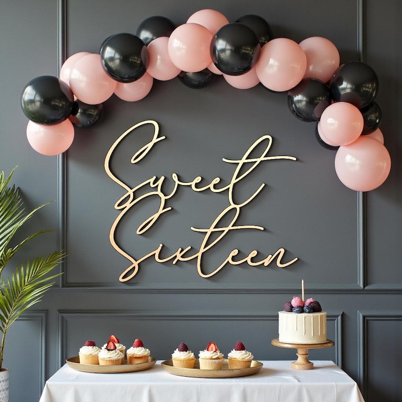 Puede incluir: Escena de fiesta con un letrero de madera "Sweet Sixteen", un arco de globos negros y rosas, cupcakes y un pastel peque&ntilde;o. El pastel est&aacute; decorado con bayas y una sola vela. El tel&oacute;n de fondo es una pared gris oscuro.