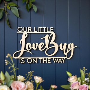 May include: A wooden sign with the words "OUR LITTLE LoveBug IS ON THE WAY" in a cursive font. The sign is light-colored and set against a dark blue background. Floral arrangements with pink and white roses and greenery frame the sign.