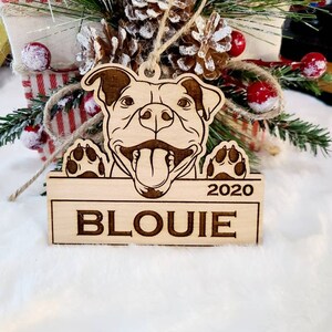 K&ouml;nnte beinhalten: Ein h&ouml;lzernes Hundeornament mit dem eingravierten Namen "BLOUIE" und dem Jahr "2020". Das Ornament zeigt ein l&auml;chelndes Hunde-Gesicht mit herausgestreckter Zunge und Pfoten. Aufgeh&auml;ngt mit Bindfaden.