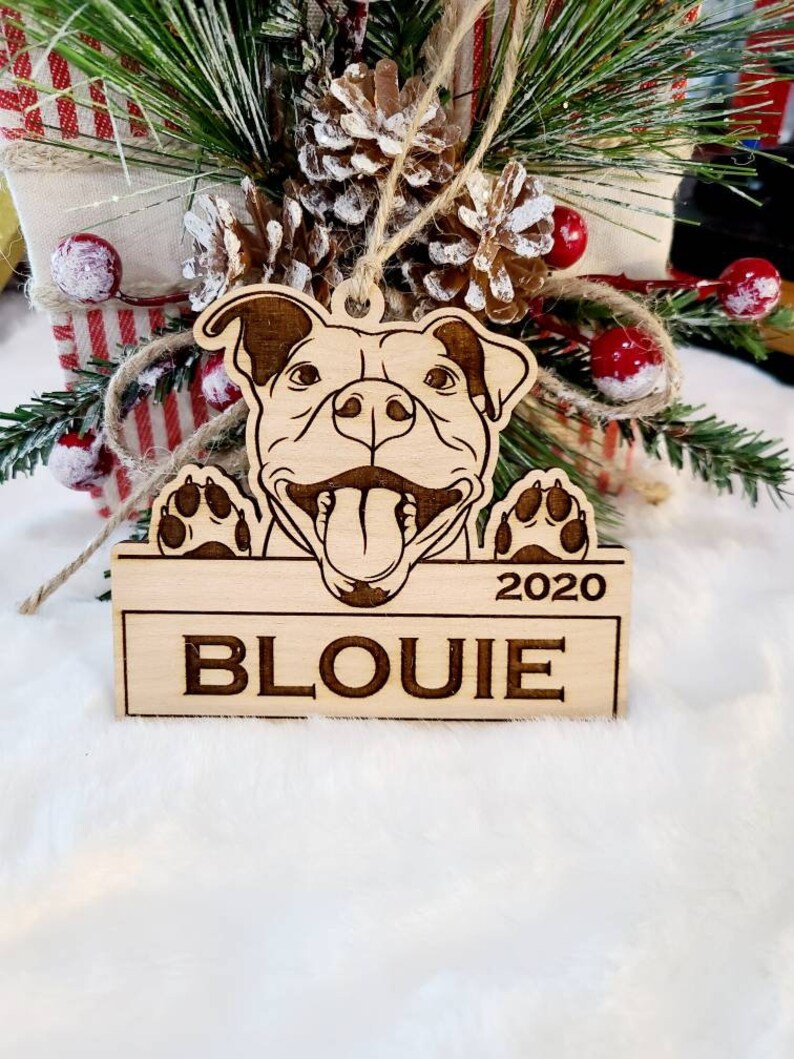 K&ouml;nnte beinhalten: Ein h&ouml;lzernes Hundeornament mit dem eingravierten Namen "BLOUIE" und dem Jahr "2020". Das Ornament zeigt ein l&auml;chelndes Hunde-Gesicht mit herausgestreckter Zunge und Pfoten. Aufgeh&auml;ngt mit Bindfaden.