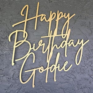 Puede incluir: Adorno para tarta de madera dorada con las palabras "Happy Birthday Goldie" en fuente cursiva. El adorno se encuentra sobre un fondo gris oscuro texturizado. Ideal para celebraciones de cumplea&ntilde;os.