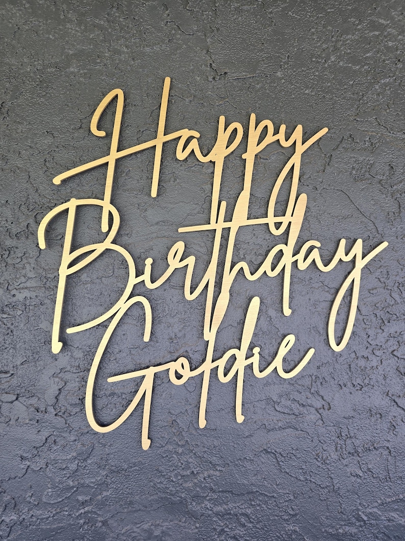 Puede incluir: Adorno para tarta de madera dorada con las palabras "Happy Birthday Goldie" en fuente cursiva. El adorno se encuentra sobre un fondo gris oscuro texturizado. Ideal para celebraciones de cumplea&ntilde;os.