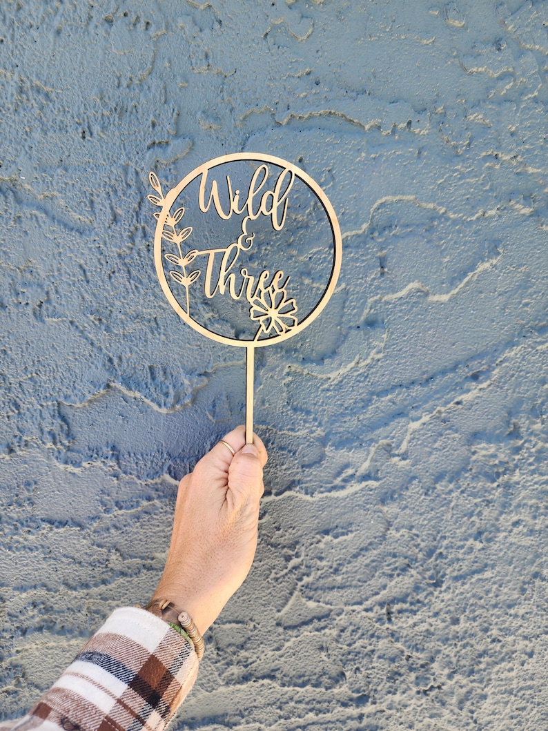 May include: Wooden cake topper with a circle design and the words "Wild & Three" in a cursive font. The circle is decorated with small floral designs.