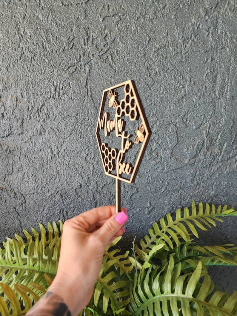 May include: Wooden cake topper shaped like a hexagon with the text "Mama to bee" and three small bees. The hexagon is decorated with honeycomb patterns.
