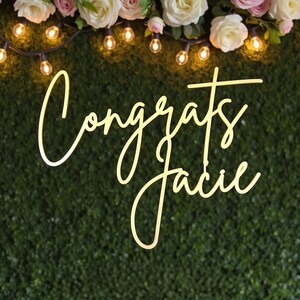 Puede incluir: Un letrero decorativo con la palabra "Congrats" y el nombre "Jacie" en fuente cursiva. El letrero est&aacute; iluminado con un brillo c&aacute;lido sobre un fondo verde. La parte superior de la imagen presenta luces de cadena y arreglos florales.