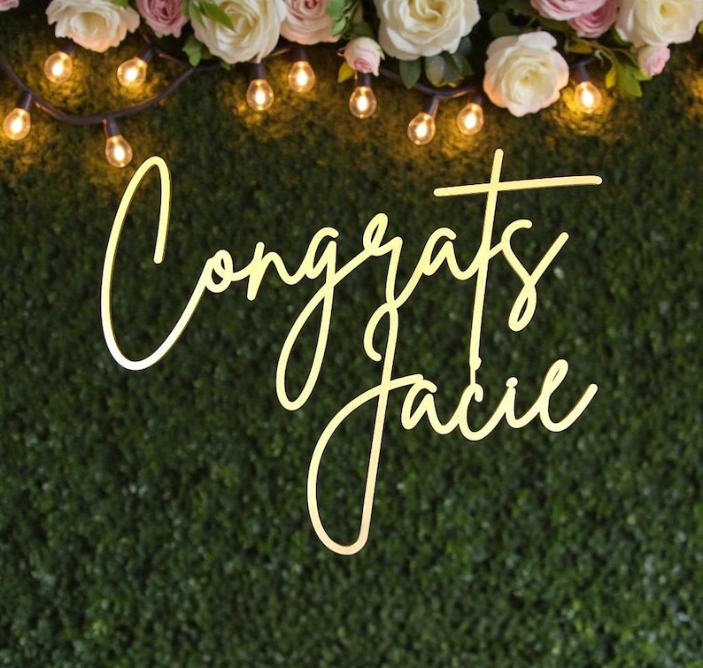 Puede incluir: Un letrero decorativo con la palabra "Congrats" y el nombre "Jacie" en fuente cursiva. El letrero est&aacute; iluminado con un brillo c&aacute;lido sobre un fondo verde. La parte superior de la imagen presenta luces de cadena y arreglos florales.