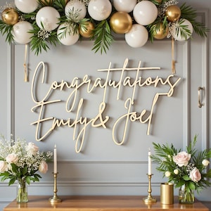 Op de afbeelding: Houten bord met de woorden "Congratulations Emily & Joel" in sierlijke letters. Het bord is bevestigd aan een grijze muur, versierd met witte en gouden ornamenten, groen en bloemstukken.