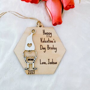 May include: Wooden ornament shaped like a gnome holding a heart with the text "XOXO". The ornament is decorated with the text "Happy Valentine's Day Brinley Love, Joshua 2021".