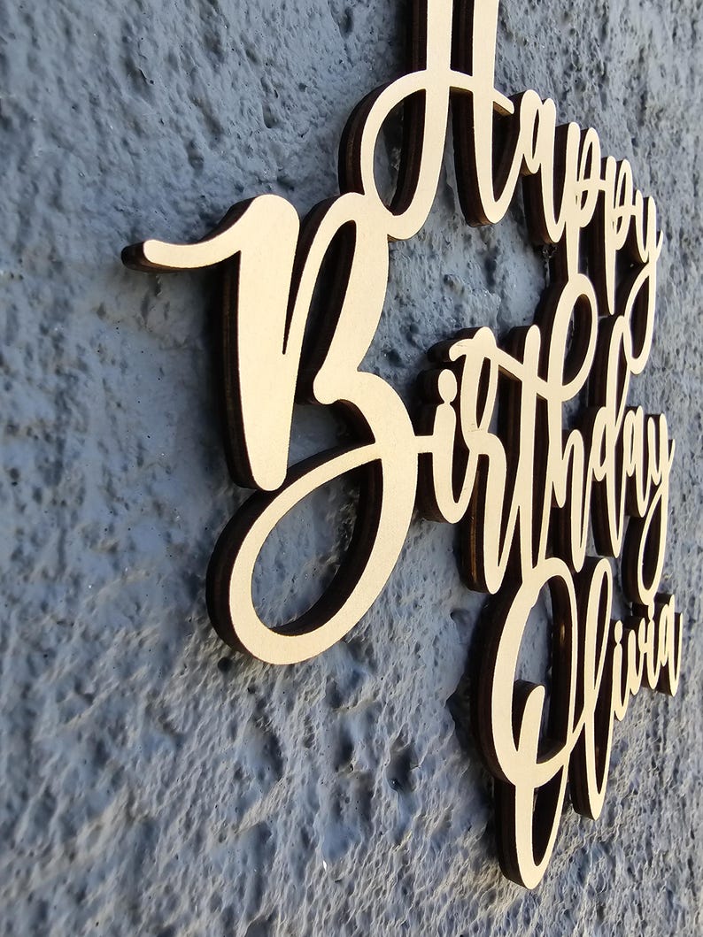 K&ouml;nnte beinhalten: Ein Holzschild mit dem Schriftzug "Happy Birthday Olivia" in einer Schreibschrift. Das Schild hat eine hellbraune Farbe mit dunkelbraunen Kanten und ist an einer strukturierten grauen Wand befestigt.