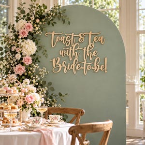 Puede incluir: Un fondo arqueado de color verde salvia presenta el texto de madera recortada Toast & Tea with the Bride-to-be! Un arreglo floral de rosas rosas y hortensias blancas se sitúa junto a una bandeja de niveles sobre una mesa blanca.