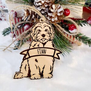 Op de afbeelding: Houten hondenornament met de naam "Finn" op een banner. Het lichtbruine ornament heeft donkerbruine ge&euml;tste details. De hond zit met zijn tong uit. Het hangt voor een feestelijk kerstarrangement.