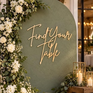 May include: An arched olive green sign with the gold script text Find Your Table, decorated with a cascading arrangement of white roses and green foliage on the left side, and two glass candle lanterns with lit white pillar candles.