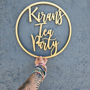 May include: A gold wooden circle with the text "Kiran's Tea Party" written in cursive.