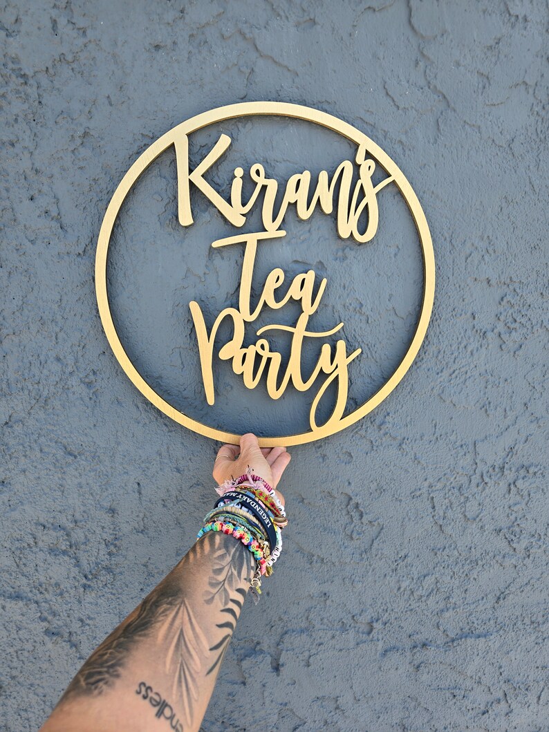 May include: A gold wooden circle with the text "Kiran's Tea Party" written in cursive.