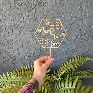 May include: Wooden hexagonal cake topper with the text "Mama to Bee" and two bees. The topper is on a stick and is held by a hand.
