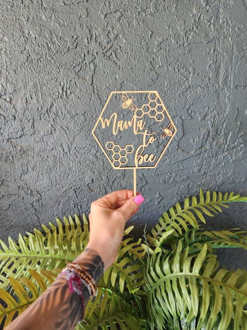 May include: Wooden hexagonal cake topper with the text "Mama to Bee" and two bees. The topper is on a stick and is held by a hand.