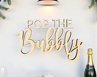 Puede incluir: Un letrero de madera con las palabras "POP THE BUBBLY" en una fuente cursiva está montado en una pared blanca. Una botella de champán, una copa de champán y un pequeño soporte blanco para pasteles con dos pasteles están sobre una mesa debajo del letrero.