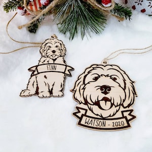 Op de afbeelding: Twee houten hondenornamenten. E&eacute;n is een volledig lichaamsbeeld van een hond met de naam "Finn" op een banner. De andere is het gezicht van een hond met de naam "Watson - 2020" op een banner. Beide ornamenten hebben een natuurlijke houten afwerking en zijn geregen met touw.
