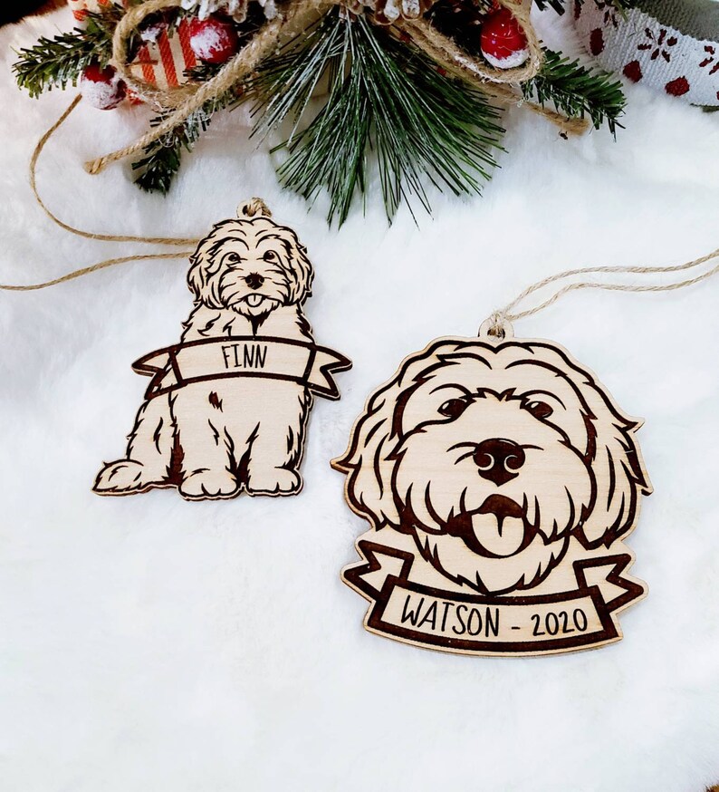 Op de afbeelding: Twee houten hondenornamenten. E&eacute;n is een volledig lichaamsbeeld van een hond met de naam "Finn" op een banner. De andere is het gezicht van een hond met de naam "Watson - 2020" op een banner. Beide ornamenten hebben een natuurlijke houten afwerking en zijn geregen met touw.