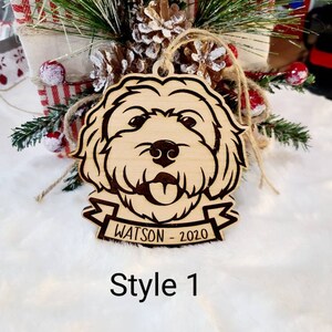 Op de afbeelding: Houten hondenornament met een gegraveerd gezicht, een banner met de tekst "WATSON - 2020" en een juten touwtje. Het ornament is gelabeld "Style 1". De achtergrond bevat feestelijk groen en rode en witte bessen.