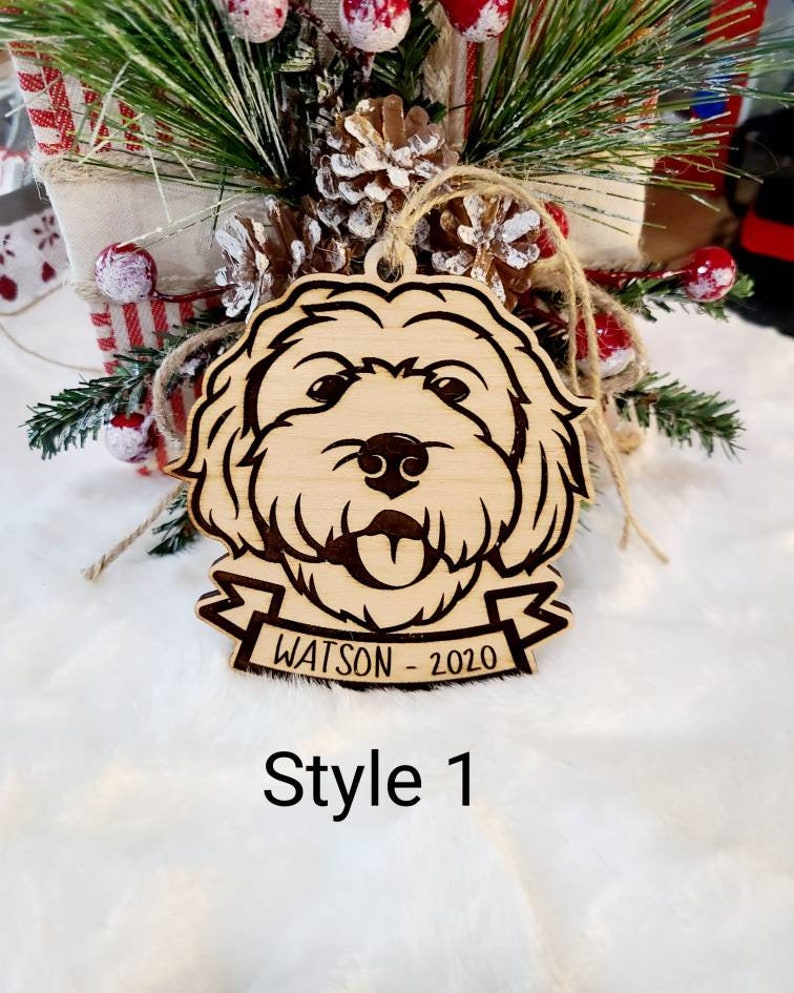 Op de afbeelding: Houten hondenornament met een gegraveerd gezicht, een banner met de tekst "WATSON - 2020" en een juten touwtje. Het ornament is gelabeld "Style 1". De achtergrond bevat feestelijk groen en rode en witte bessen.