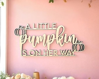 May include: Wooden sign with the text "A Little Pumpkin Is On Her Way" in a cursive font, with pumpkin illustrations. The sign is displayed on a pink wall, with pumpkins and floral arrangements.