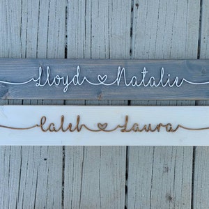 May include: Two wooden signs with names written in wire. The top sign is gray with white wire and says "Lloyd & Natalie". The bottom sign is white with copper wire and says "Caleb & Laura".