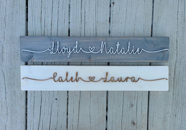 May include: Two wooden signs with names written in wire. The top sign is gray with white wire and says "Lloyd & Natalie". The bottom sign is white with copper wire and says "Caleb & Laura".