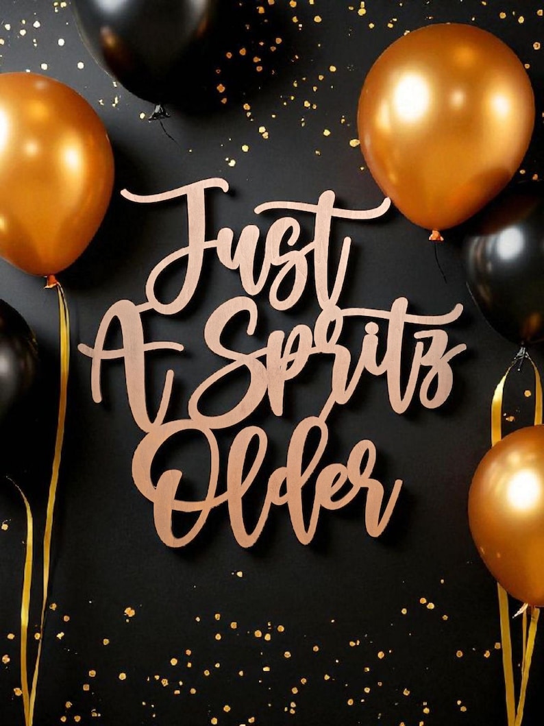 Puede incluir: Un fondo negro con confeti dorado y globos dorados y negros. Un letrero de madera con la inscripci&oacute;n "Just A Spritz Older". Los globos est&aacute;n atados con cintas doradas.