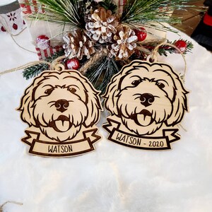 Op de afbeelding: Twee houten kerstversieringen in de vorm van een hond. Elk ornament heeft een gedetailleerd lasergesneden ontwerp van een hondengezicht met zijn tong uitgestoken. E&eacute;n ornament heeft de naam "WATSON" op een banner, en de andere zegt "WATSON - 2020".