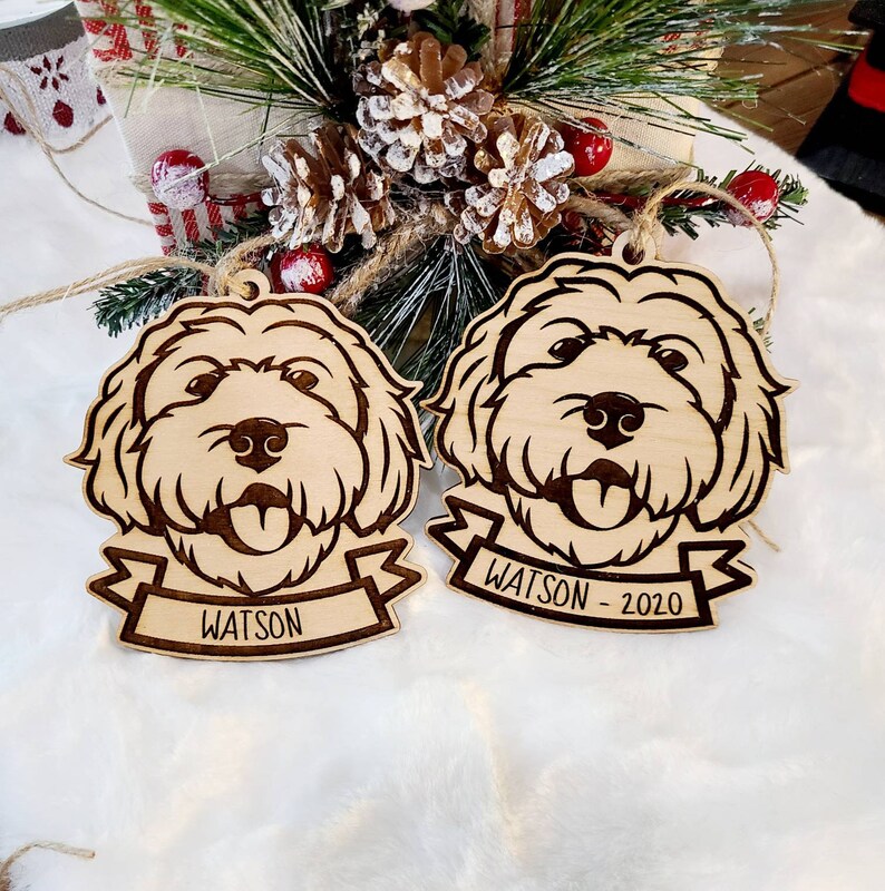 Op de afbeelding: Twee houten kerstversieringen in de vorm van een hond. Elk ornament heeft een gedetailleerd lasergesneden ontwerp van een hondengezicht met zijn tong uitgestoken. E&eacute;n ornament heeft de naam "WATSON" op een banner, en de andere zegt "WATSON - 2020".
