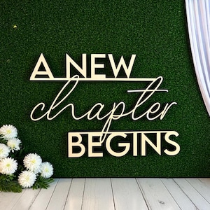 May include: Wooden sign with the words "A NEW chapter BEGINS" in a decorative font. The sign is set against a green textured backdrop, with white flowers and a white curtain on either side. The sign is perfect for a wedding or event.