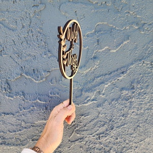 May include: Wooden cake topper with a circle design and the text "Wild & Three" in a brown wood finish. The topper has a small floral design and a stick for inserting into a cake.