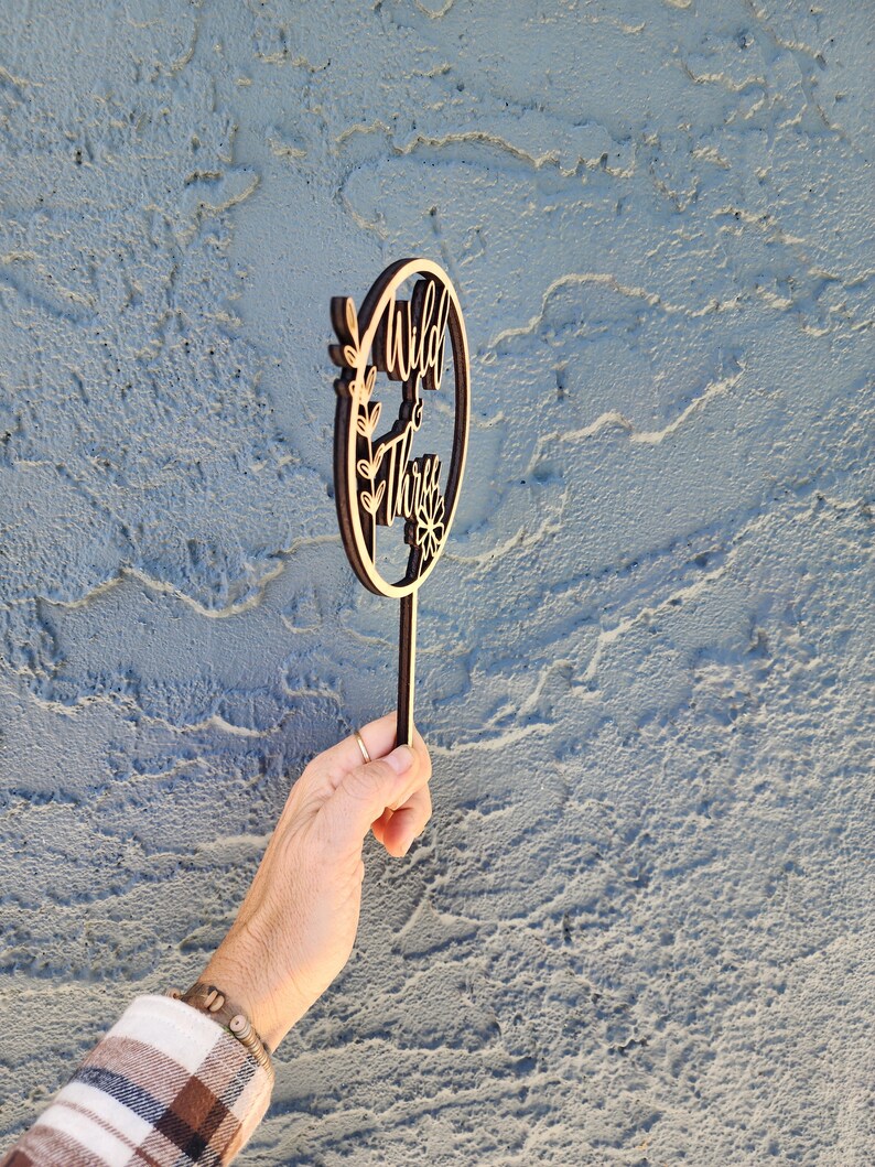 May include: Wooden cake topper with a circle design and the text "Wild & Three" in a brown wood finish. The topper has a small floral design and a stick for inserting into a cake.