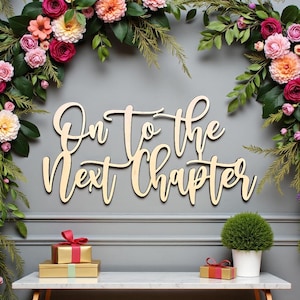 Puede incluir: Letrero de madera con la frase "On To the Next Chapter" en escritura cursiva. El letrero está rodeado de arreglos florales con flores rosas, naranjas y blancas. Dos regalos envueltos y una planta en maceta están sobre una mesa blanca.