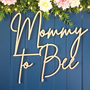 Puede incluir: Letrero de madera con las palabras "Mommy to Bee" en fuente cursiva, sobre un fondo azul. El letrero est&aacute; decorado con vegetaci&oacute;n, luces de hadas y flores rosas y blancas, ideal para un baby shower.
