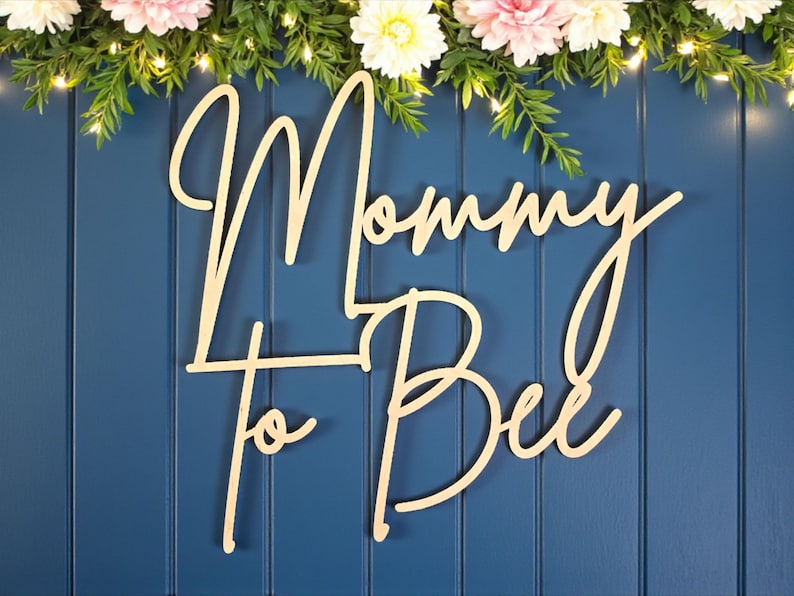 Puede incluir: Letrero de madera con las palabras "Mommy to Bee" en fuente cursiva, sobre un fondo azul. El letrero est&aacute; decorado con vegetaci&oacute;n, luces de hadas y flores rosas y blancas, ideal para un baby shower.