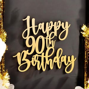 Puede incluir: Adorno dorado de pastel de madera con las palabras "Happy 90th Birthday" en una elegante escritura. El adorno est&aacute; sobre un fondo oscuro, con serpentinas doradas y flores blancas en las esquinas.