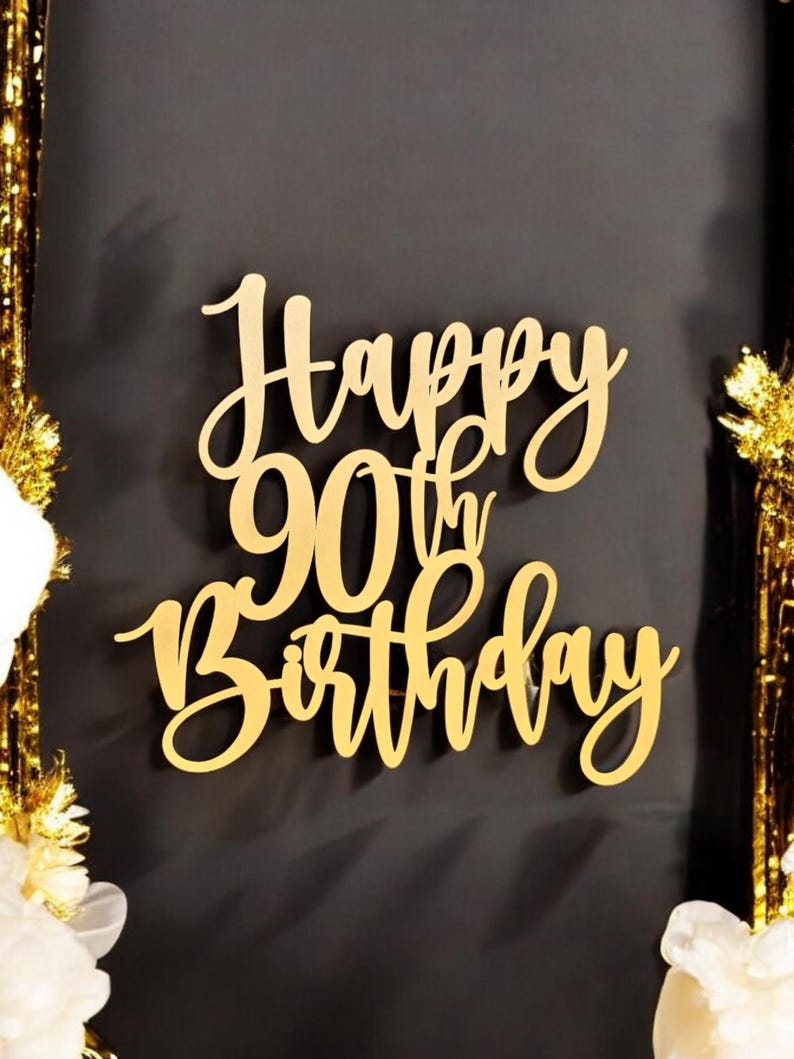 Puede incluir: Adorno dorado de pastel de madera con las palabras "Happy 90th Birthday" en una elegante escritura. El adorno est&aacute; sobre un fondo oscuro, con serpentinas doradas y flores blancas en las esquinas.