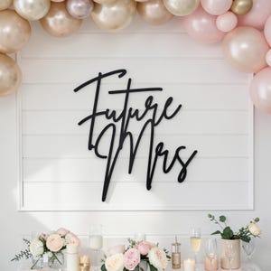 May include: A black wooden sign with the words "Future Mrs" in elegant script is mounted on a white paneled wall. The sign is the focal point, surrounded by a balloon arch and floral arrangements, creating a celebratory atmosphere.