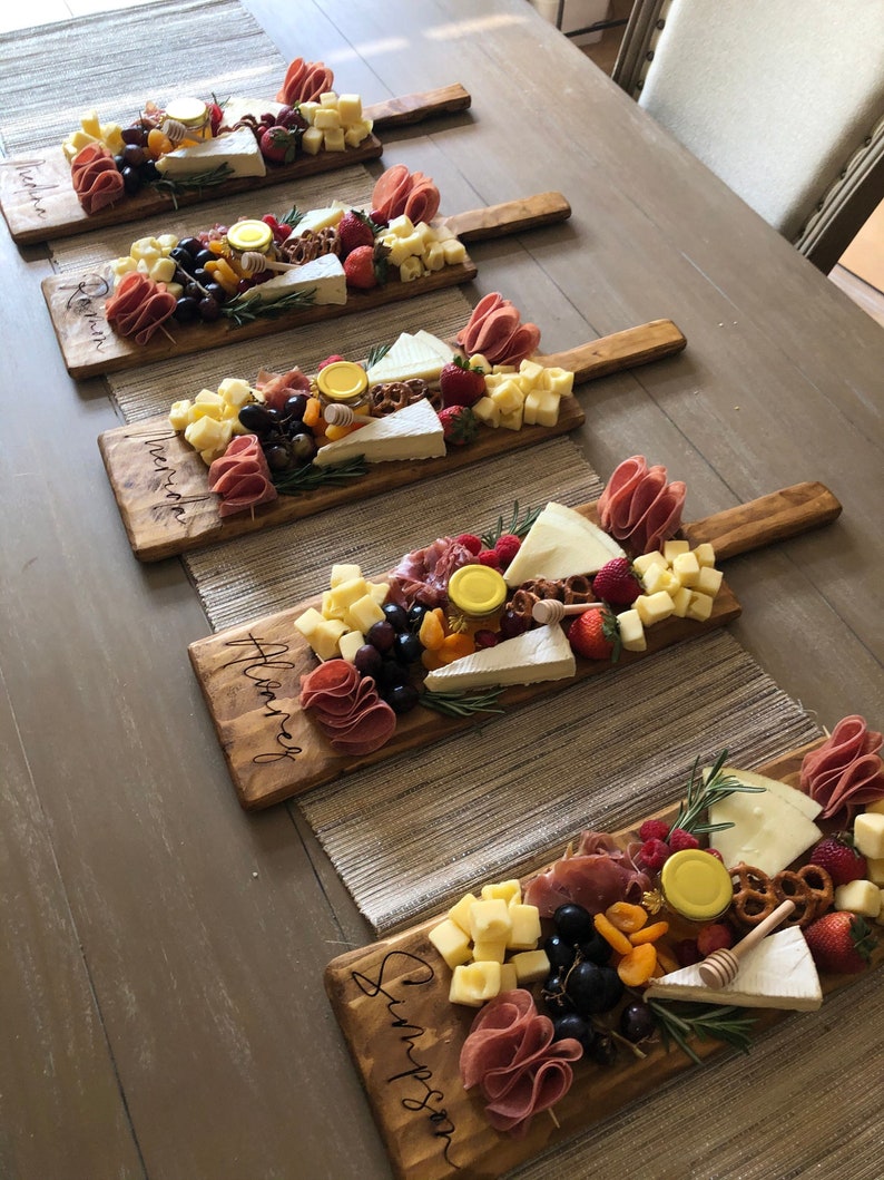 Puede incluir: Tablas de charcuter&iacute;a de madera dispuestas sobre una mesa, cada una cargada con una variedad de quesos, carnes, frutas y miel. Las tablas tienen asas y est&aacute;n grabadas con nombres. Tambi&eacute;n se ven fresas, uvas y pretzels.