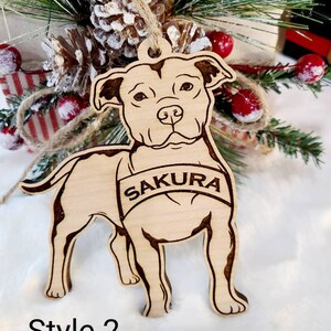 K&ouml;nnte beinhalten: Hellbrauner Holzschmuck in Form eines Hundes mit dem Namen "Sakura" auf einem Banner eingraviert. Der Schmuck hat dunkelbraune, ge&auml;tzte Details. Der Text "Style 2" befindet sich darunter. Er h&auml;ngt vor einer Weihnachtsdekoration.