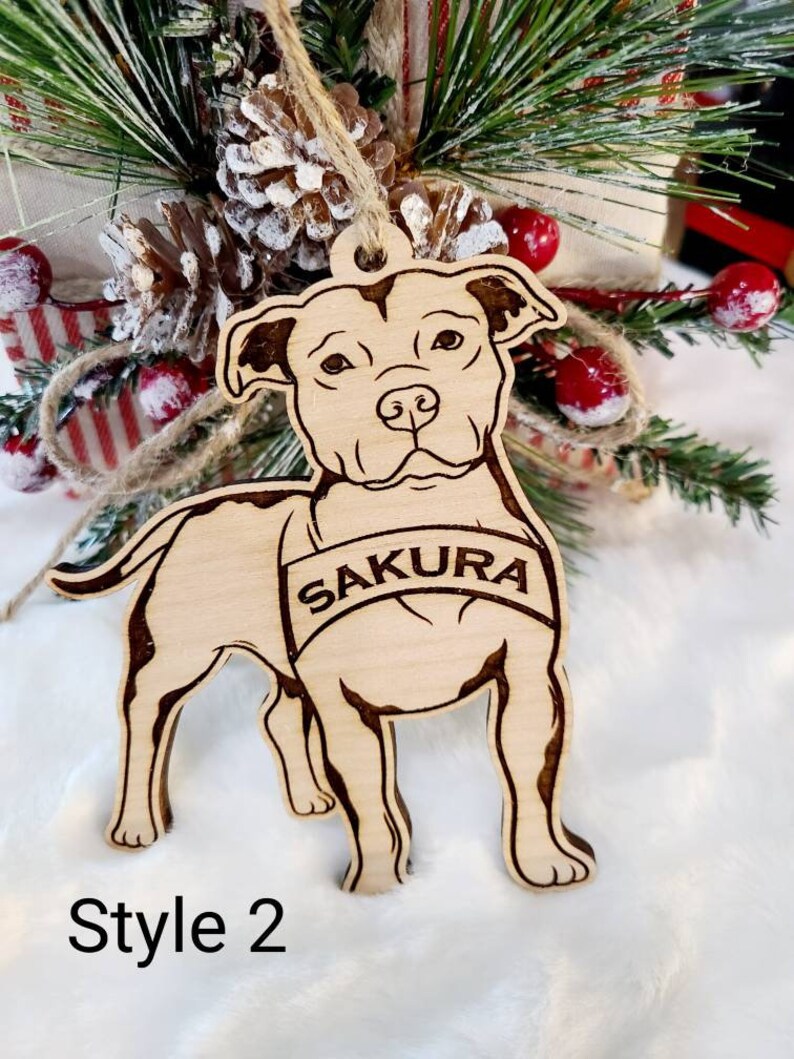 K&ouml;nnte beinhalten: Hellbrauner Holzschmuck in Form eines Hundes mit dem Namen "Sakura" auf einem Banner eingraviert. Der Schmuck hat dunkelbraune, ge&auml;tzte Details. Der Text "Style 2" befindet sich darunter. Er h&auml;ngt vor einer Weihnachtsdekoration.