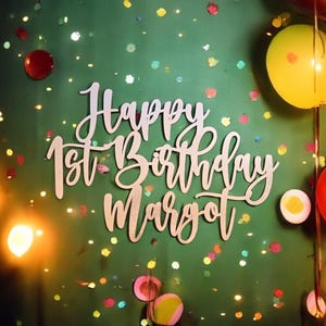 Puede incluir: Un letrero de madera que dice "Happy 1st Birthday Margot" en escritura cursiva. El letrero est&aacute; sobre un fondo verde decorado con confeti, globos y luces, creando un ambiente festivo.