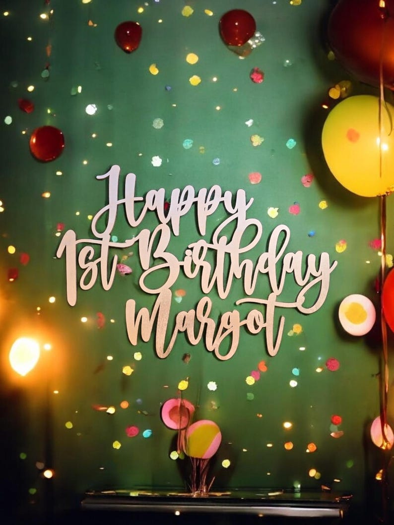 Puede incluir: Un letrero de madera que dice "Happy 1st Birthday Margot" en escritura cursiva. El letrero est&aacute; sobre un fondo verde decorado con confeti, globos y luces, creando un ambiente festivo.