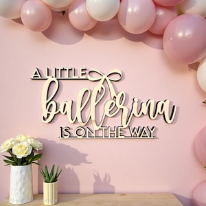 May include: Wooden sign with the text "A Little Ballerina Is On The Way" in a cursive font. The sign is light brown and mounted on a pink wall, with pink and white balloons in the background. A vase of white flowers and a small succulent are in the foreground.