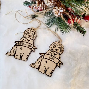 Op de afbeelding: Twee houten hondenornamenten met de namen "Maggie - 2020" en "Finn" op banners. De lichtbruine ornamenten hebben donkerbruine contouren en touwhangers. Feestelijk groen en dennenappels op de achtergrond.