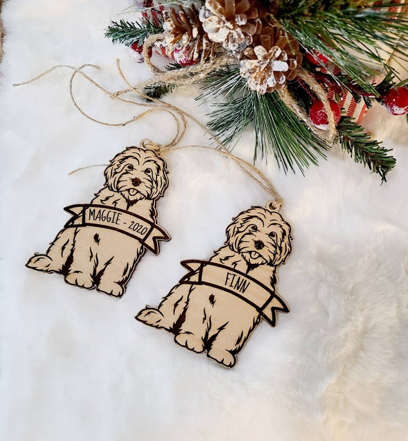 Op de afbeelding: Twee houten hondenornamenten met de namen "Maggie - 2020" en "Finn" op banners. De lichtbruine ornamenten hebben donkerbruine contouren en touwhangers. Feestelijk groen en dennenappels op de achtergrond.