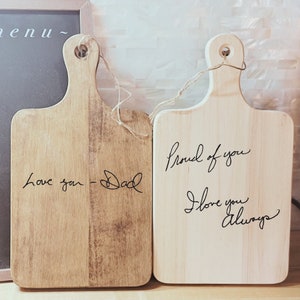 May include: Two wooden cutting boards with handwritten messages. The first board says "Love you - Dad". The second board says "Proud of you I love you always".